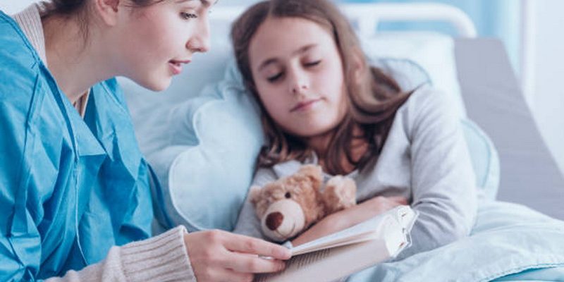 Older sister reading book to unhealthy girl with plush toy in private clinic